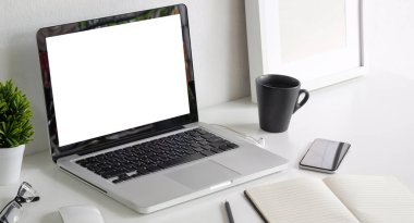 Computer laptop with blank display, smart phone, coffee cup, frame and houseplant on white table at modern workspace.