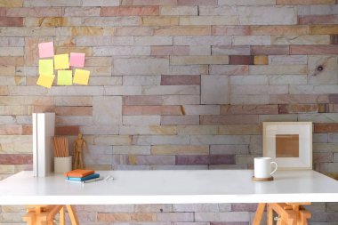 Designer workplace, pencil holder, books, picture frame and coffee cup on white table with brick wall.