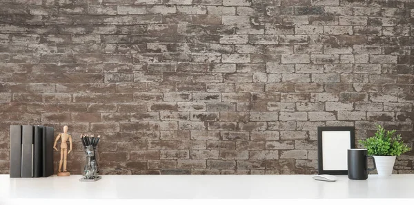 Creative workplace, picture frame, books, coffee cup and houseplant on white table with brick wall.