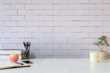 Kitaplar, akıllı telefon, elma, kahve fincanı ve tuğla duvarlı beyaz masa üzerindeki saksı bitkisi..