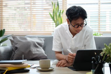 Evde bilgisayar tabletiyle çalışan odaklanmış Asyalı genç adam..