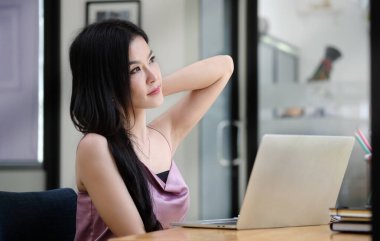 Tatmin olmuş Asyalı genç bir kadın iş yerinde dinleniyor ve ofisin penceresinden bakıyor..