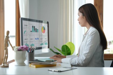 Konsantre kadın iş adamı parlak bir ofiste oturup bilgisayar ekranında finansal grafiği analiz ediyor..