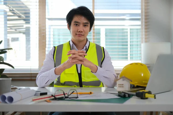 身穿正装 头戴硬礼帽 头戴滚动式蓝图的年轻亚洲工程师坐在工作场所 对着相机微笑 — 图库照片