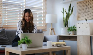 Oturma odasında kanepede oturan ve dizüstü bilgisayar kullanan mutlu genç bir kadın..