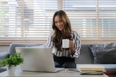 Güzel genç bir kadın parlak oturma odasında kanepede oturuyor ve dizüstü bilgisayar kullanıyor..
