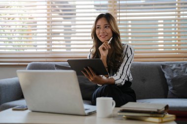 Dizüstü bilgisayarın karşısında oturan ve dijital tablet kullanan gülümseyen serbest çalışan genç bir kadın..