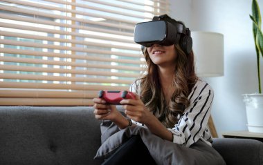 Sanal gerçeklik kulaklığı takan ve elinde joysticklerle evde video oyunları oynayan heyecanlı bir kadın..
