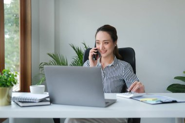 Çekici Asyalı genç iş kadını modern ofiste oturuyor ve cep telefonuyla konuşuyor..