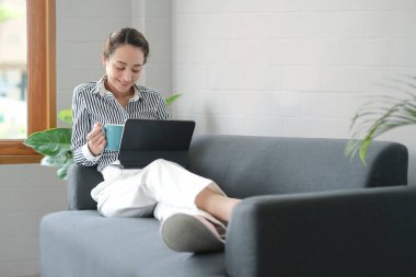 Gülümseyen milenyum kadını rahat koltukta otururken kahve içiyor ve dijital tablette haberler okuyor..