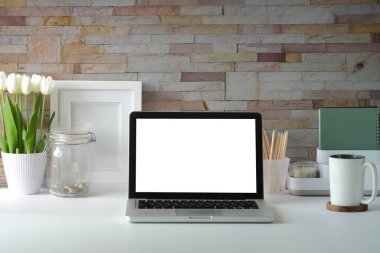 Boş ekranlı dizüstü bilgisayar, saksı, kahve fincanı ve resim çerçevesi beyaz masada..