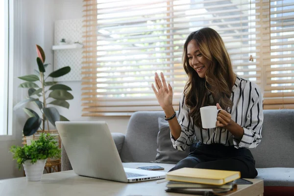 Gülümseyen genç bir kadın, evinde dizüstü bilgisayarla video görüşmesi yaparken arkadaşına el sallıyor..