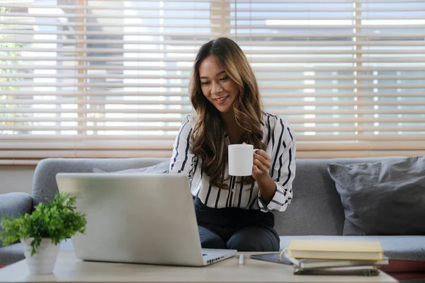 Güzel genç bir kadın parlak oturma odasında kanepede oturuyor ve dizüstü bilgisayar kullanıyor..