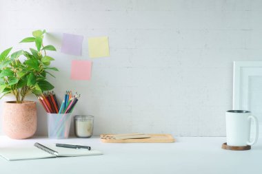 Resim çerçeveli, kahve fincanı, saksı bitkisi ve beyaz masadaki malzemeleri olan şık bir iş yeri. Boşluğu kopyala.