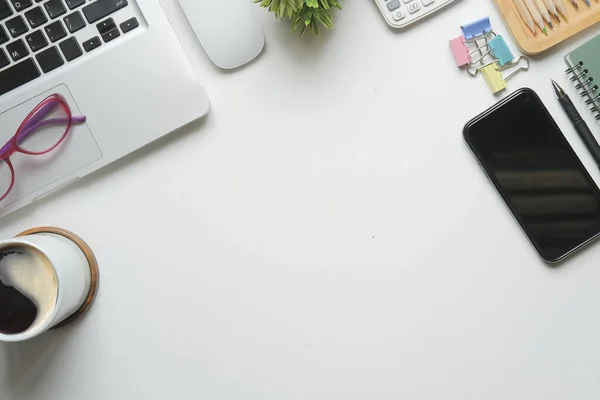 Akıllı telefonu, dizüstü bilgisayarı, kahve fincanı ve bardakları olan beyaz ofis masası. Üst manzara, düz uzanma.