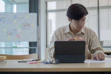 Bilgisayar tableti kullanan ve ofis masasındaki mali raporları kontrol eden odaklanmış genç işadamı..