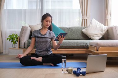 Smiling young woman using digital tablet while sitting on exercise mat at home. Sport, fitness and healthy lifestyle concept.