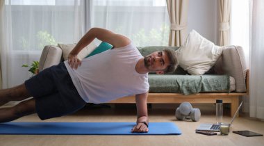 Aktif adam aydınlık oturma odasında paspas üzerinde yan tahta egzersizi yapıyor. Spor, spor ve sağlıklı yaşam tarzı konsepti.