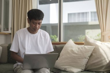 Smiling handsome asian man sitting on floor in living room and using laptop computer.	