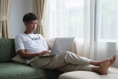 Oturma odasında kanepede oturan ve dizüstü bilgisayar kullanan mutlu genç adam..