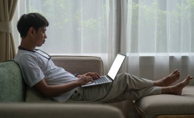 Rahat genç adam evdeki rahat koltukta oturuyor ve dizüstü bilgisayar kullanıyor..