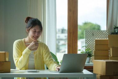 Asyalı kadın satıcı dizüstü bilgisayar kullanıyor, internetten ev ofisindeki siparişleri kontrol ediyor. Başlangıçta küçük işletme, çevrimiçi satış, E-ticaret kavramı.