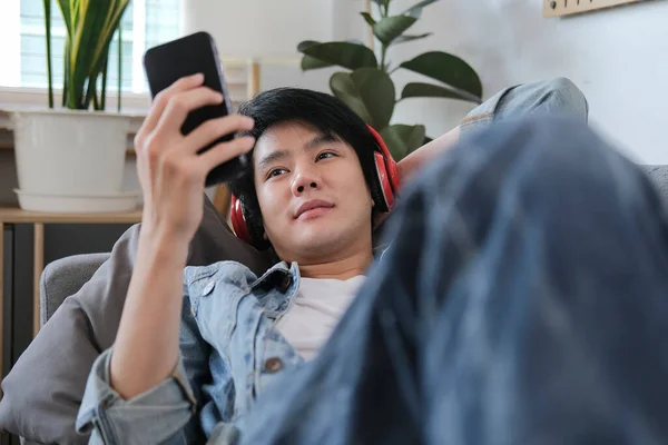 Випадковий Молодий Азіатський Чоловік Бездротовими Навушниками Лежачи Дивані Використовуючи Смартфон — стокове фото