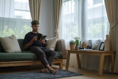 Genç adamın parlak oturma odasındaki koltukta otururken akıllı telefon kullanmasının tam boy görüntüsü..