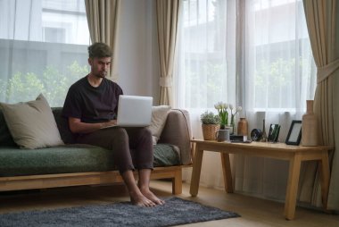 Gülümseyen yakışıklı Asyalı adam oturma odasında oturmuş dizüstü bilgisayar kullanıyor..