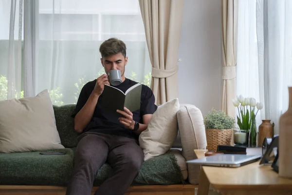 Olumlu Genç Adam Kitap Okuyor Evdeki Kanepede Otururken Kahve Içiyor — Stok fotoğraf