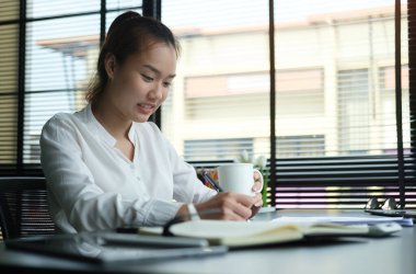 Asyalı kadın ofis çalışanı kahve içiyor ve modern ofiste çalışıyor..