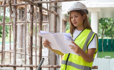 İnşaat alanının planlarını inceleyen güvenlik ceketi ve kasklı kadın mimar..