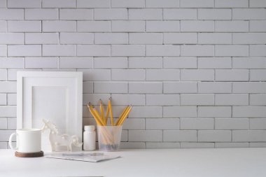 Boş resim çerçevesi, kahve fincanı, kitap ve kırtasiyesi beyaz masada, tuğla duvara dayalı rahat bir iş yeri. Metnin için boşluk kopyala.