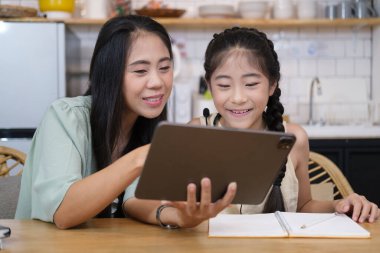 Asian mother helping her daughter doing homework with digital tablet at home.	