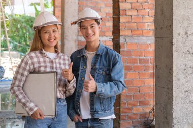 İnşaat alanında güvenlik kaskı takan ve kameraya başparmağını gösteren kendine güvenen mimarlar..