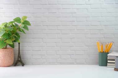 Saksı bitkisi, kalem tutacağı ve beyaz masada tuğladan duvara yaslanmış kitapları olan minimum işyeri. Metnin için boşluk kopyala.