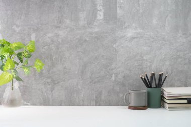 Kahve fincanı, kitap, kalem tutacağı ve tavan arası duvarına karşı beyaz masa üzerinde saksı bitkisi olan şık bir iş yeri. Metnin için boşluk kopyala.