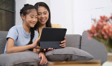 Asyalı anne ve tatlı küçük kızı evdeki kanepede dijital tablet kullanıyorlar. Hafta sonu aile eğlencesi..