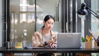 Genç Asyalı kadın yaratıcı tasarımcı modern işyerinde bilgisayar laptopuyla çalışıyor..