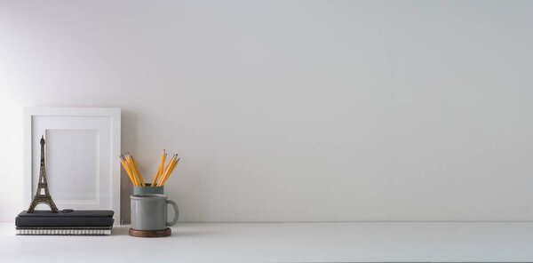 Minimal workplace with empty picture frame, book, coffee cup and pencil holder on white table. Copy space for your text.