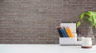 Kahve fincanı, kırtasiye malzemesi ve beyaz masa üzerinde tuğla duvarlı ev bitkisi olan şık bir iş yeri. Merkez ofis masası.