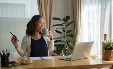Oturma odasında dizüstü bilgisayarın önünde oturan ve sıcak kahve içen orta yaşlı, rahat bir kadın..