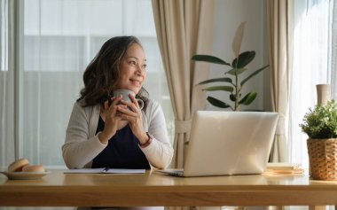 Kafası rahat orta yaşlı bir kadın kahve fincanı tutuyor ve başka tarafa bakıyor. Stressiz, huzurlu bir ruh halinin keyfini çıkarıyor. Oturma odasında yalnız başına..