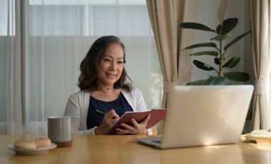 Zarif orta yaşlı kadın uzaktan kumandayla evden çalışırken dizüstü bilgisayarından online webinar izliyor..