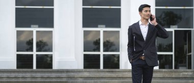 Zarif genç işadamı cep telefonuyla konuşuyor ve modern ofis bölgesinde dikilirken düşünceli görünüyor. .