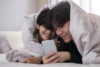 Genç eşcinsel çift battaniyenin altında yatıyorlar ve birlikte akıllı telefon kullanıyorlar..