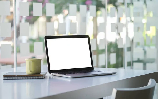 Laptop computer with white screen, books and coffee cup on wooden desk near window.