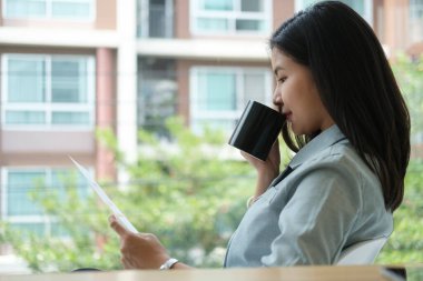 Yandan bakan iş kadını sıcak kahve içiyor ve modern ofisin büyük penceresinin yanında finansal belgeler okuyor..