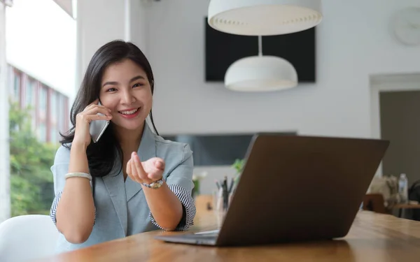 陽気なアジアの女性従業員が彼女のラップトップの前に座って携帯電話で話して — ストック写真