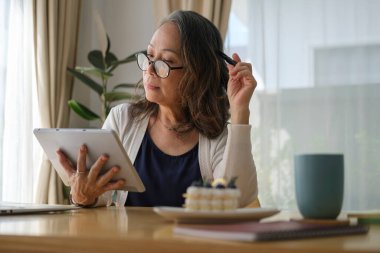 Güzel olgun bir kadın parlak oturma odasında oturuyor ve dijital tablet kullanıyor, internette sörf yapıyor, sabahları haberleri kontrol ediyor..